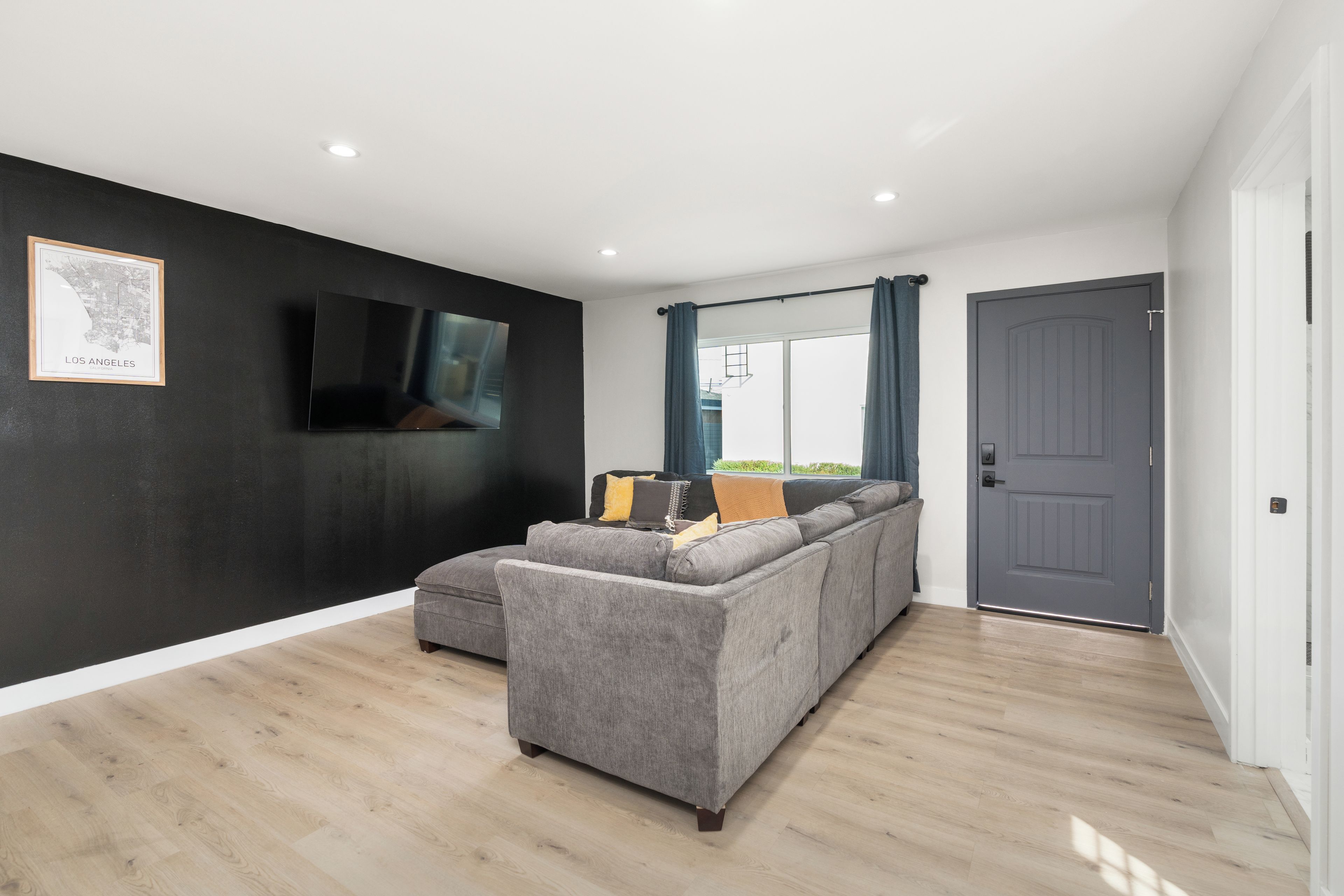 Un salon moderne avec un canapé gris, un téléviseur à écran plat fixé au mur et une grande fenêtre avec des rideaux.