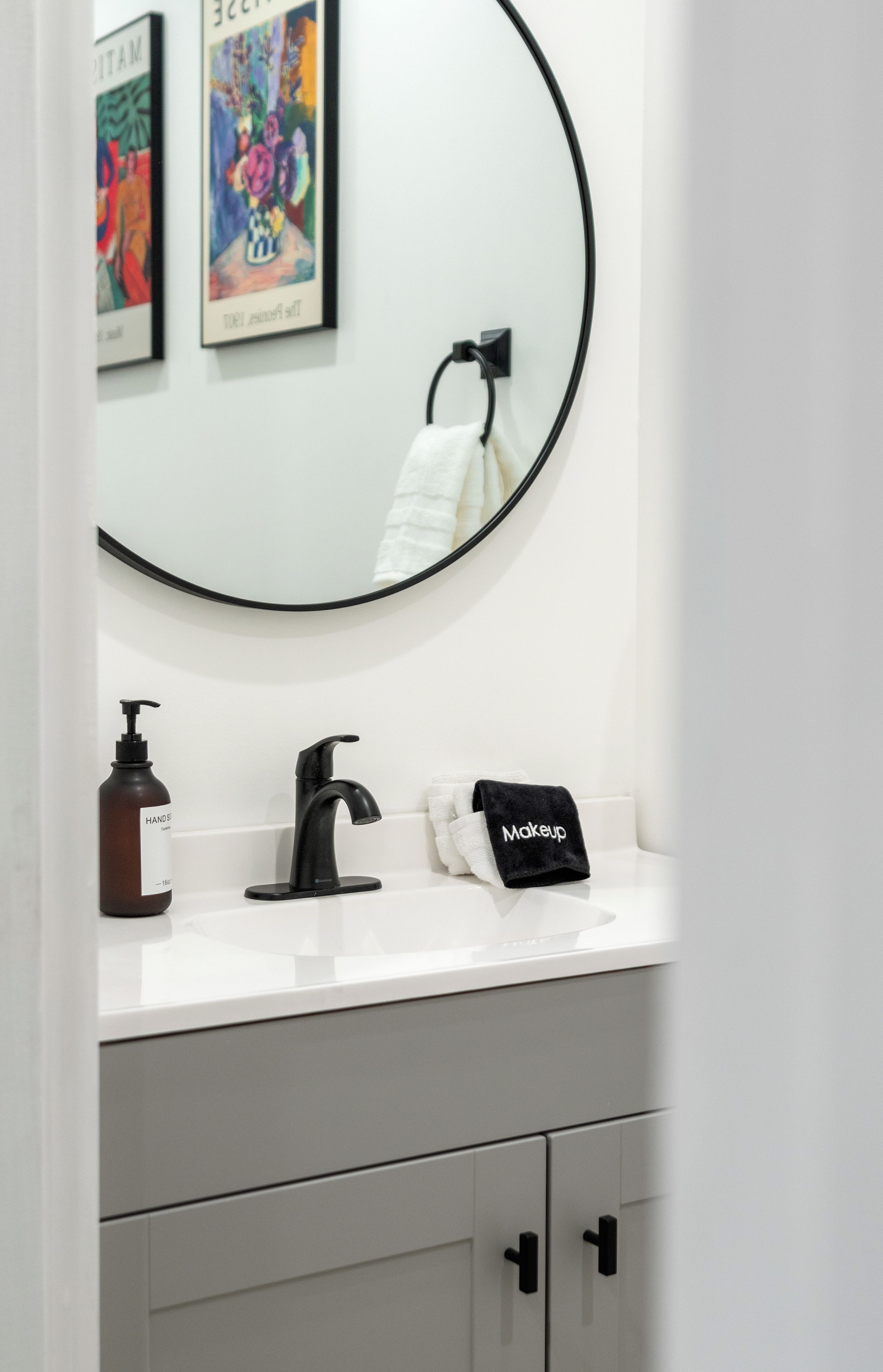 Ein Badezimmer mit einem weißen Waschbecken, einem schwarzen Wasserhahn, einer Handseifenflasche, einem schwarzen Handtuch mit dem Aufdruck 'Makeup' und einem Spiegel mit schwarzem Rahmen.