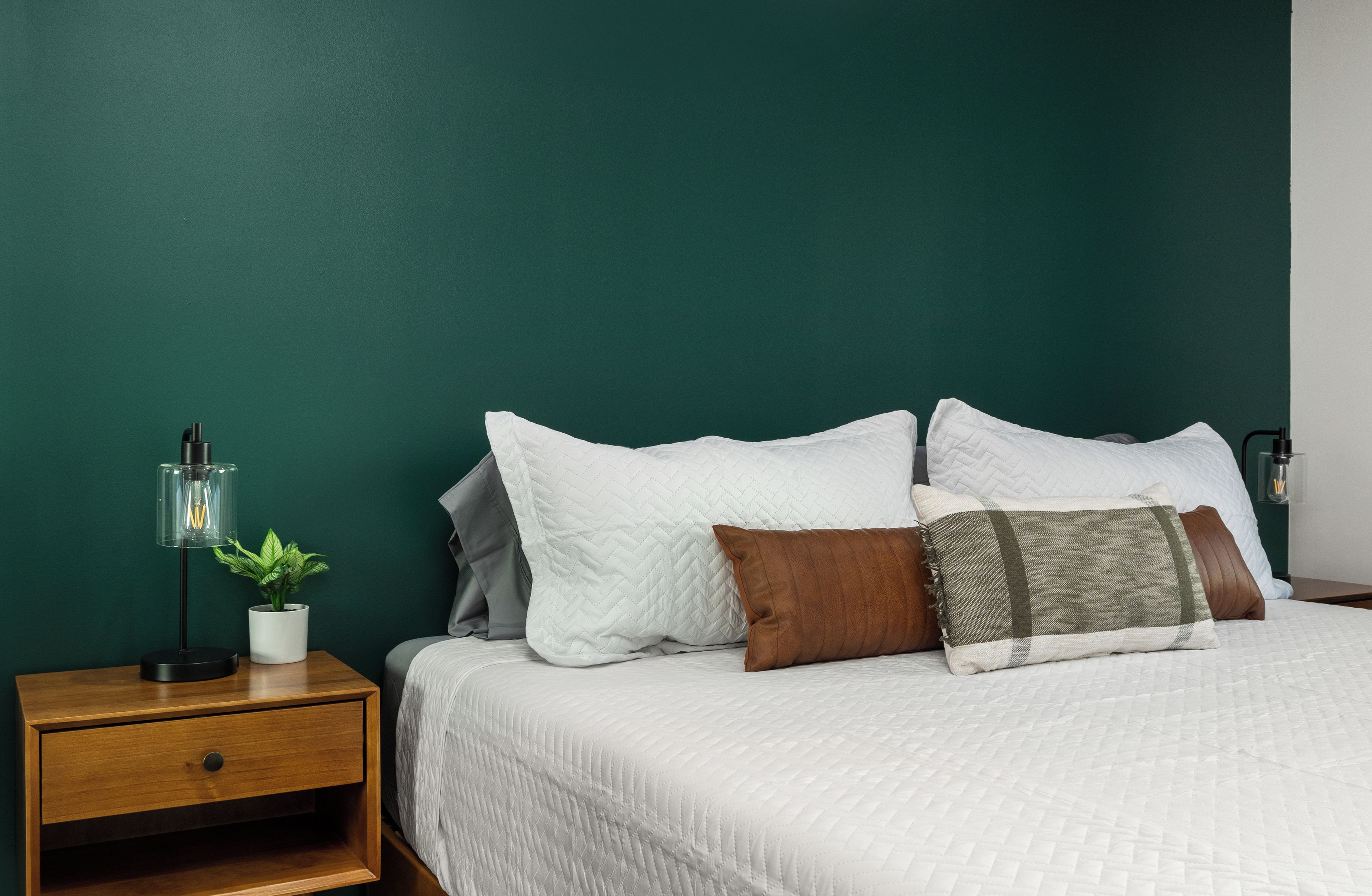 Un lit bien fait, avec du linge de lit blanc et plusieurs oreillers, une table de chevet avec une lampe et une plante en pot, et un mur vert.