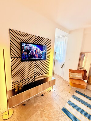 A modern living room with a flat-screen TV mounted on a geometric-patterned wall, a wooden entertainment unit below, a floor lamp, and a striped rug. - Stylish Studio in the Heart of Cannes – Central Location & Contemporary Comfort” (Cannes)