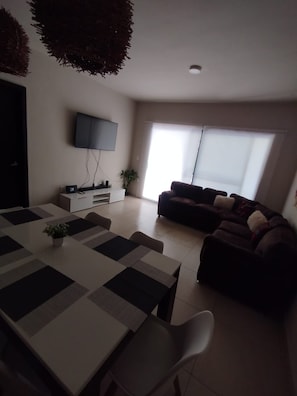 Ein modernes Wohnzimmer mit Fernseher, einer Couch, einem Esstisch mit Stühlen und einem großen Fenster.