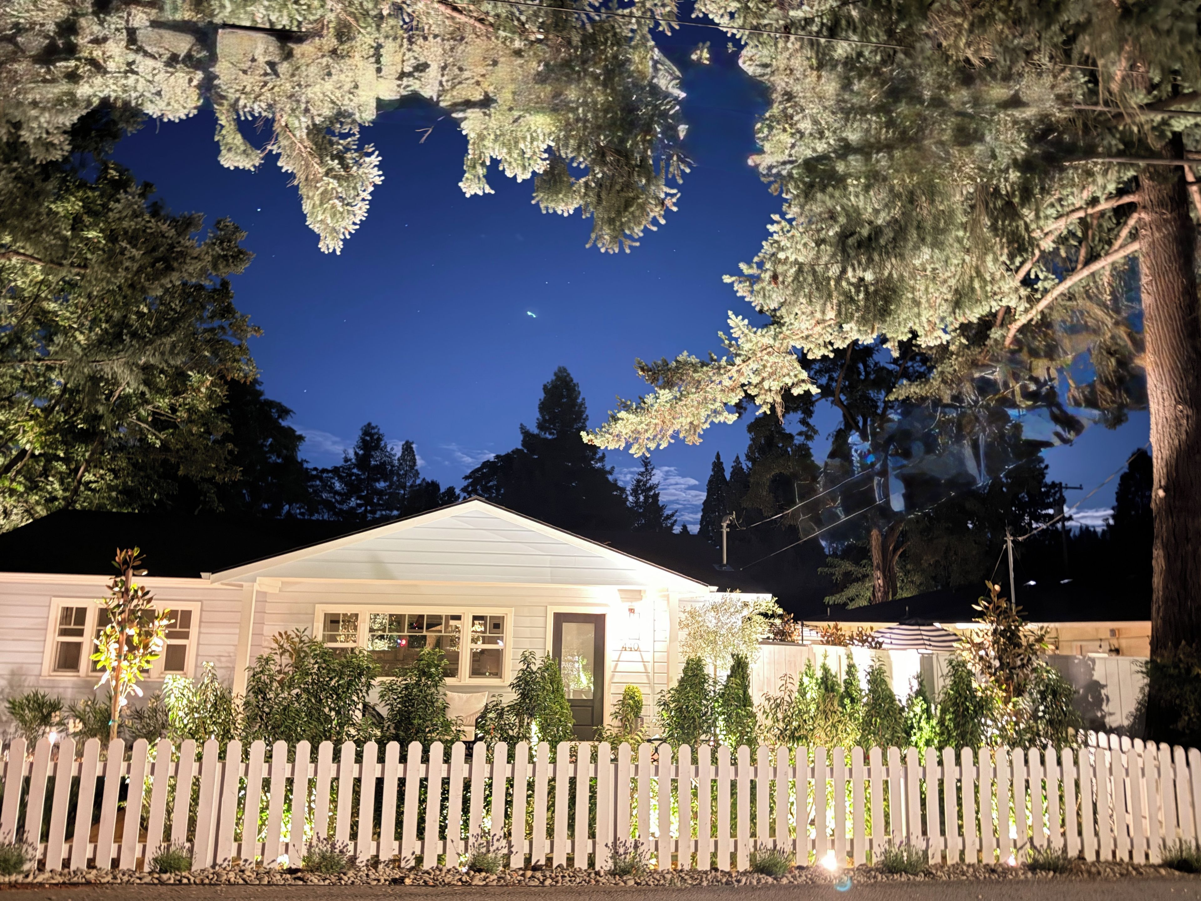 A house with a white picket fence, surrounded by trees and a clear night sky.