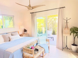 A bedroom with a bed, bedside table, lamp, potted plant, and a view of the outdoors. - Bluebird Bungalow- Renovated Mid-Century Dream in the Historic Core of J-Ville! (Jacksonville)
