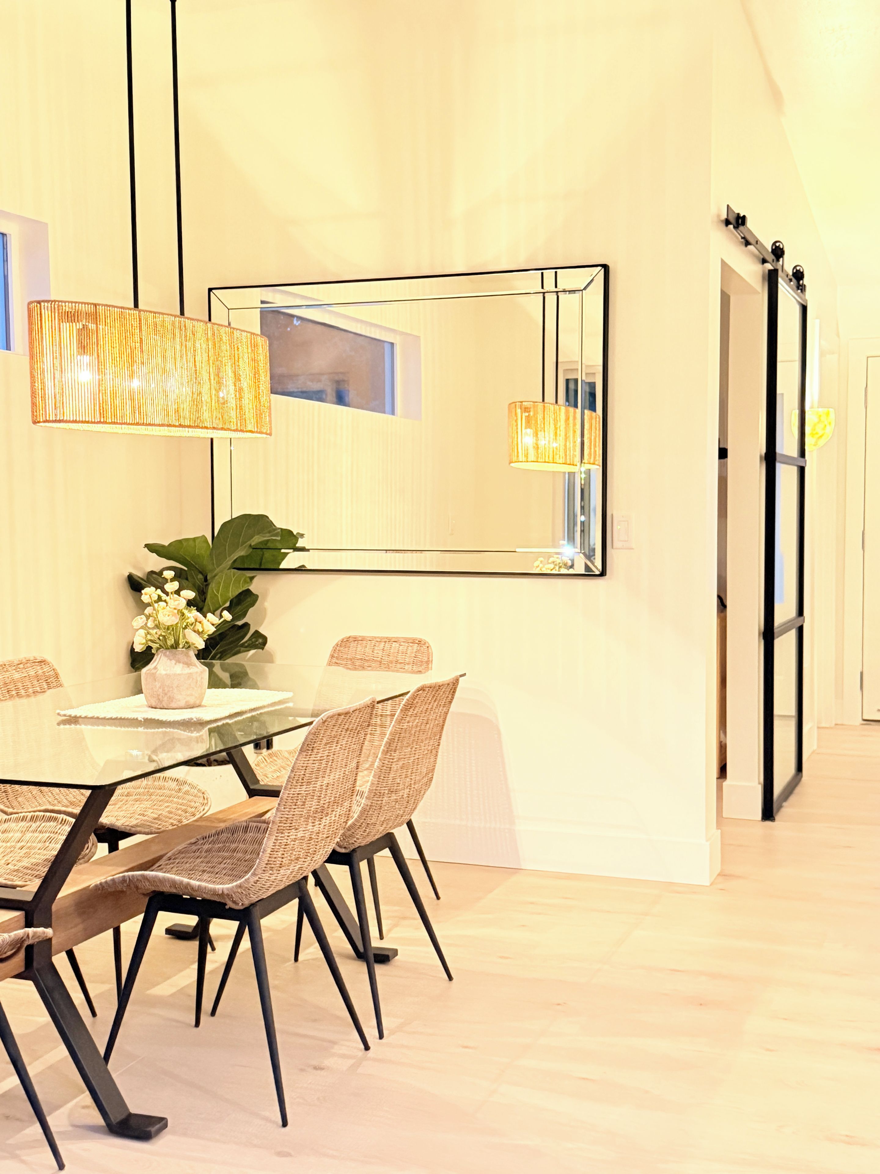 A modern dining area with a glass table, wicker chairs, and a large mirror on the wall.