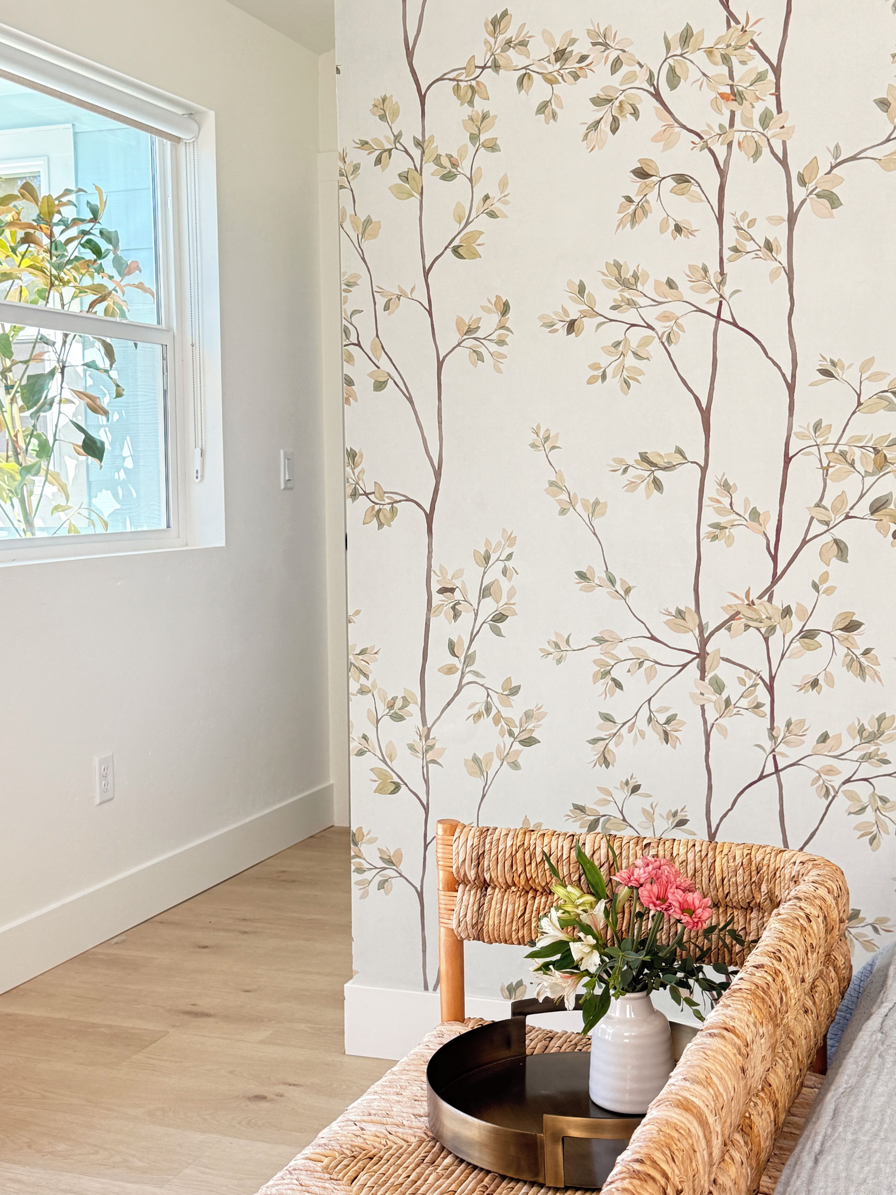 A room with a wooden floor, a white wall with a tree branch pattern, a wicker bench, and a vase with flowers.