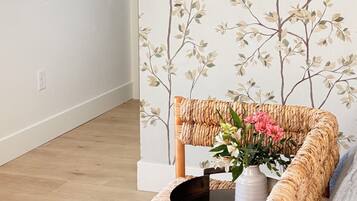 A room with a wooden floor, a white wall with a tree branch pattern, a wicker bench, and a vase with flowers.