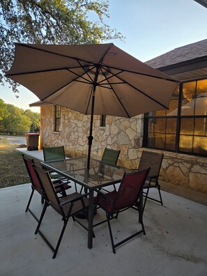 A glass-top table with four chairs and a large umbrella, set on a patio with a stone wall and a window. - Quiet oak-lined retreat in Garden Ridge - 4BR home (Garden Ridge)