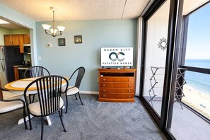 A room with a dining table and chairs, a wooden dresser, a wall-mounted TV, and a large window with a view of the beach. - Golden Sands 1611111 (Ocean City)
