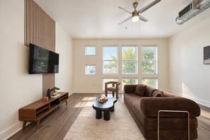 A living room with a flat-screen TV, a wooden entertainment unit, a brown sofa, a coffee table, and a ceiling fan. - Spacious 1BR | 850 Sq Ft | Design District (Dallas)