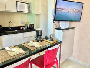 A kitchenette with a microwave, coffee maker, and a TV displaying a coastal scene. - Cozy 1-bedroom apartment with AC in beautiful São Luís (São Luís)