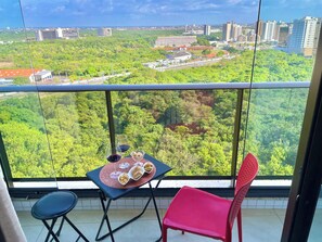A balcony with a table set for two, a red chair, and a view of a cityscape and greenery. - Cozy 1-bedroom apartment with AC in beautiful São Luís (São Luís)