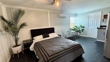 A bedroom with a bed, a ceiling fan, a potted plant, a small table, and a view of the kitchen.