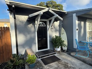 Une maison avec une porte d’entrée noire, un petit porche, une chaise bleue et une planche de surf appuyée contre le mur.