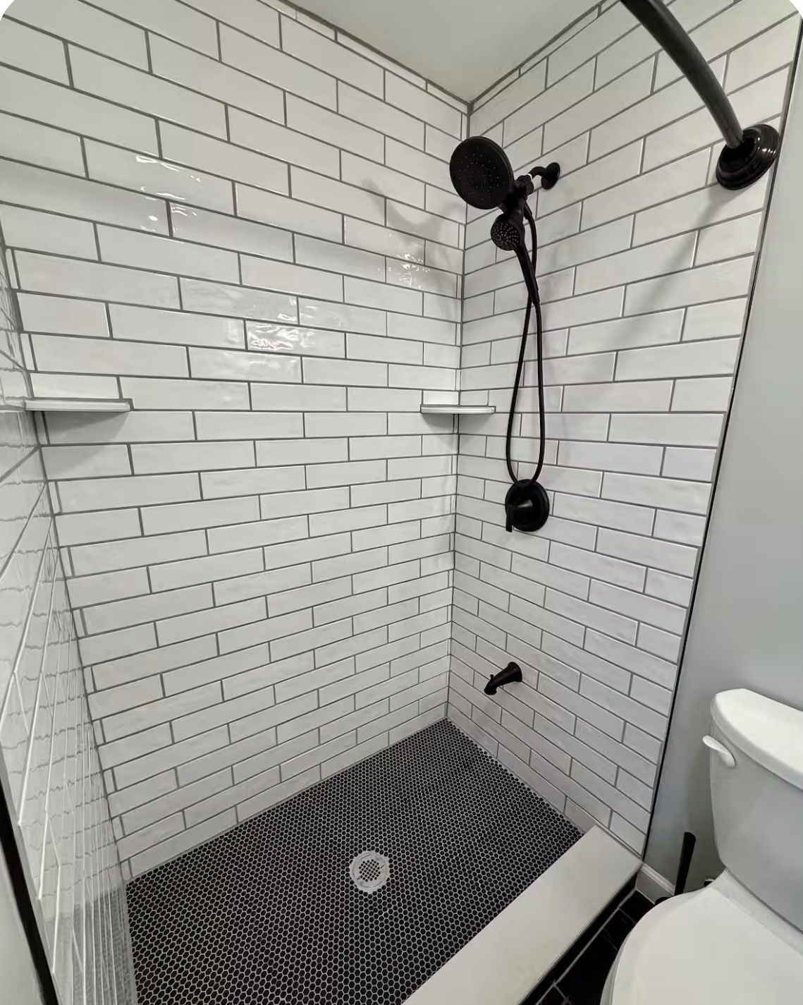 Eine Dusche mit weißen Fliesen im U-Bahn-Stil, einem schwarzen Duschkopf und einer weißen Toilette.