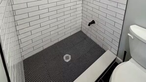 En dusch med vita, rektangulära kakelplattor, ett svart duschmunstycke och en vit toalett.