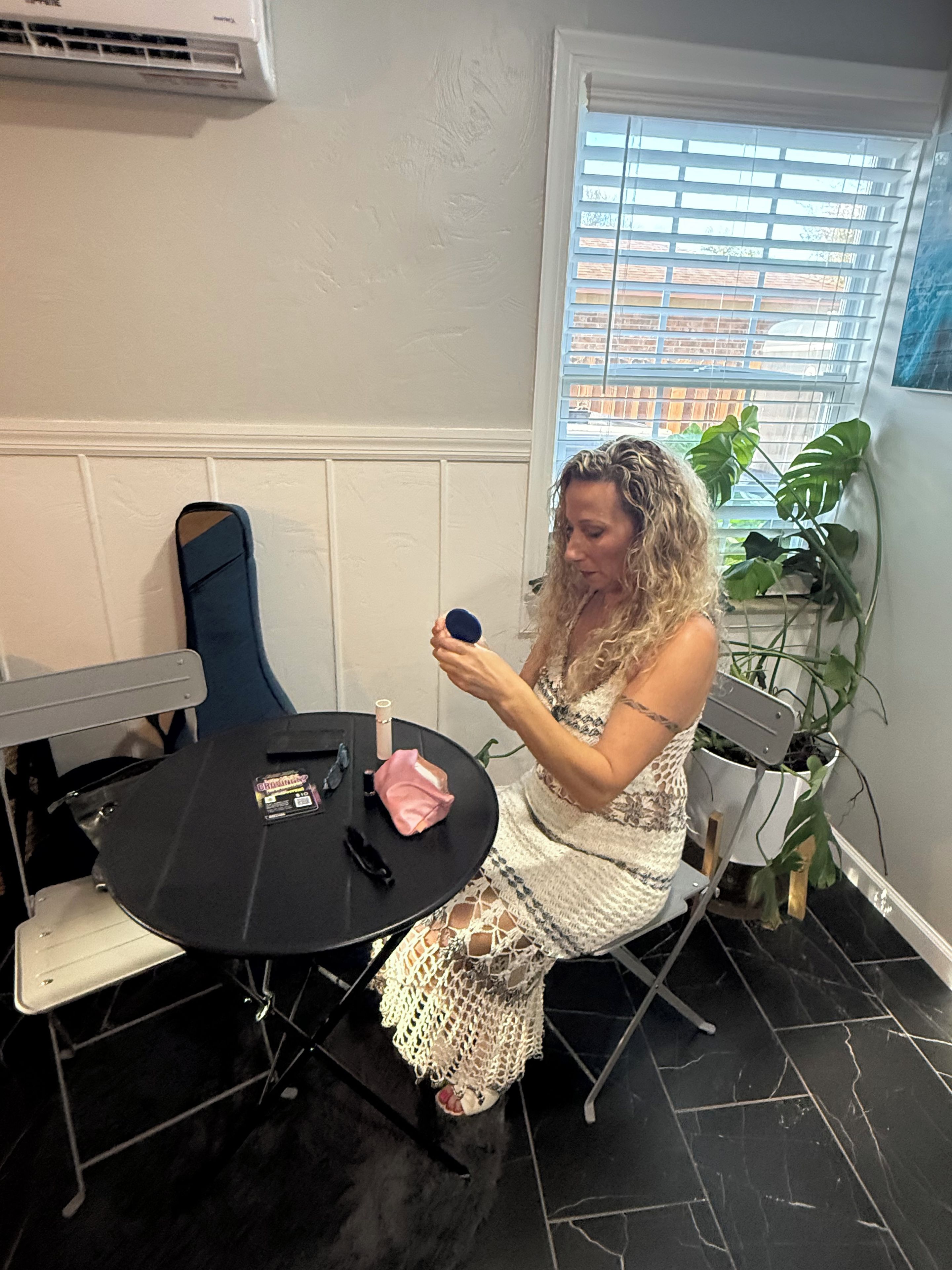 A person seated at a round table with makeup items, a chair, and a potted plant in a room with a window and an air conditioning unit.