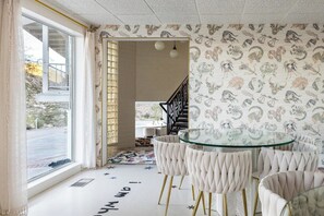 Ein Zimmer mit einem Glastisch, umgeben von ausgestatteten Sesseln, einem gemusterten Wandpapier und Blick durch ein Fenster.