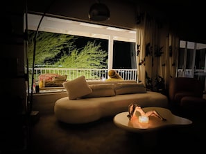 Ein schwach beleuchteter Raum mit einer Couch und einem Tisch, ein Balkon mit Pflanzen und Blick auf den Nachthimmel.