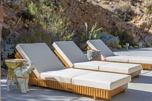 A row of sun loungers with wooden frames and white cushions, arranged on a patio with a rocky backdrop and desert vegetation. - ZAINALAND!! Mythic Desert Den!! (Twentynine Palms)