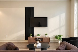 Ein modernes Wohnzimmer mit einem wandmontierten Flachbildfernseher, einem Holzkonsolentisch mit Pflanzen, einem runden Couchtisch mit Büchern und zwei Sofas.