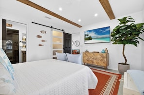 A bedroom with a bed, a wardrobe, a mirror, a plant, and a painting on the wall.