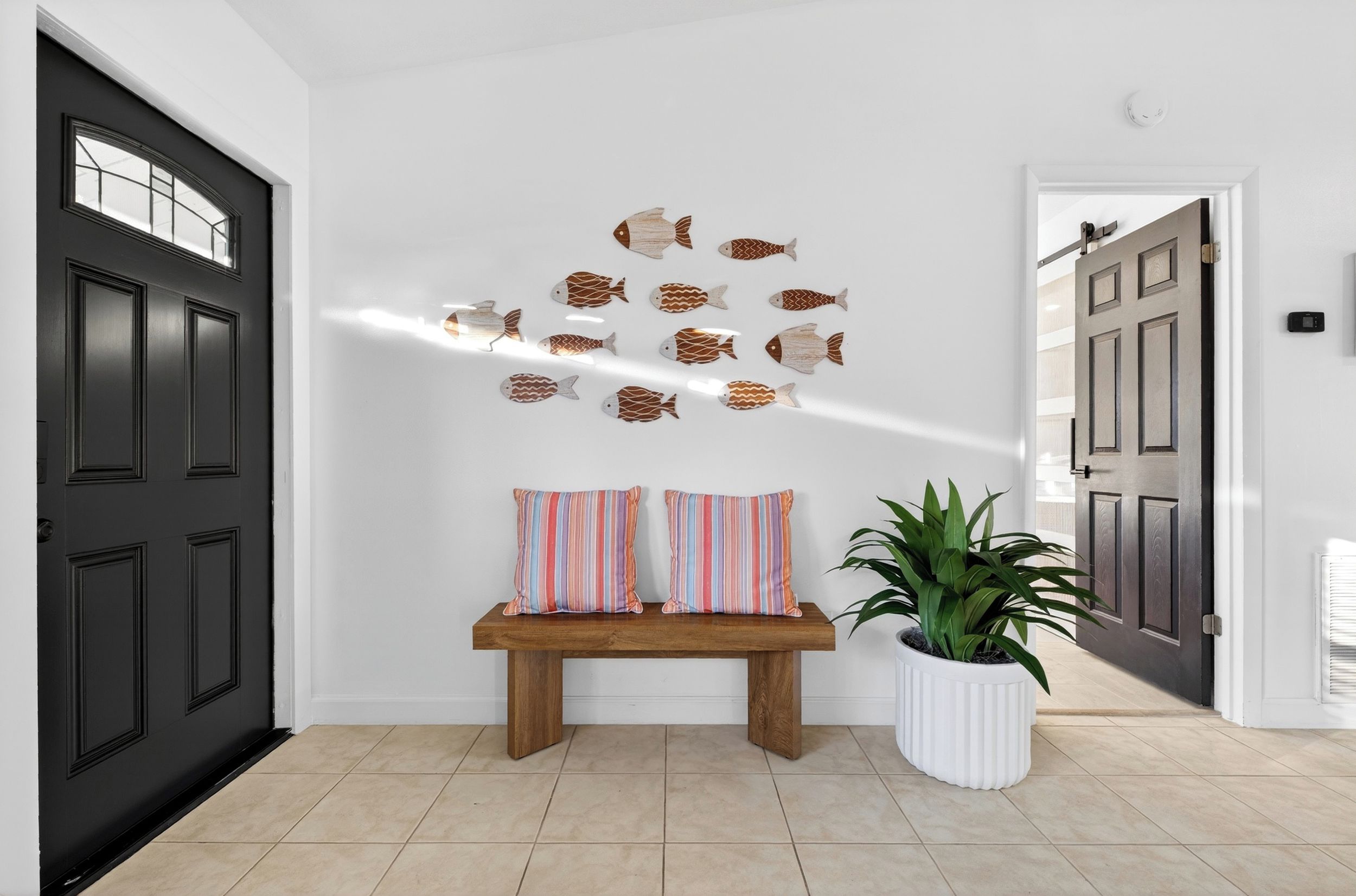 Un pasillo con una puerta negra, un banco con cojines a rayas, una decoración de pez montada en la pared y una planta en maceta.