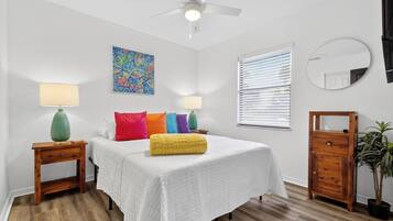 Un dormitorio con una cama, dos mesitas de noche, un ventilador de techo, una ventana con persianas y un espejo fijado a la pared, además de un cuadro de peces de colores.