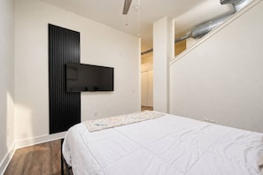 Ein Schlafzimmer mit einem Bett, einem an der Wand befestigten Fernseher und freiliegenden Lüftungsrohren.