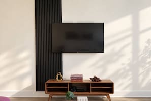 Ein modernes Wohnzimmer mit einem flachbildfernseher an der Wand, einem hölzernen Konsolentisch mit Büchern und einem dekorativen Gegenstand, sowie einer Pflanze auf dem Boden.