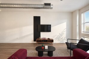 A modern living room with a flat-screen TV mounted on the wall, a wooden console table, a round coffee table, and red upholstered sofas. - Design District 1BR | Reserved Parking | Sleeps 4 (Dallas)