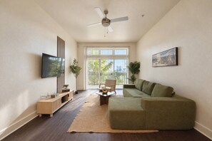 A living room with a green sofa, a TV mounted on the wall, and a ceiling fan. - Design District 2BR/2BA | Sleeps 6 | Parking (Dallas)