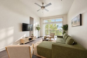Un salon moderne avec un canapé vert, un meuble TV en bois, un ventilateur de plafond et une grande fenêtre donnant sur l’extérieur.