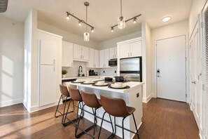 A modern kitchen with white cabinetry, a central island with bar stools, stainless steel appliances, and pendant lighting. - Modern 2BR/2BA | 3 Queen Beds | Design District (Dallas)
