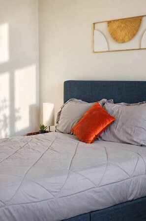 A bed with a quilted cover, a blue headboard, and a single orange pillow. - Modern 2BR/2BA | 3 Queen Beds | Design District (Dallas)