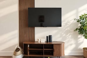 A flat-screen TV mounted on a wall above a wooden console table with a speaker and a small white object on it. - Modern 2BR/2BA | 3 Queen Beds | Design District (Dallas)