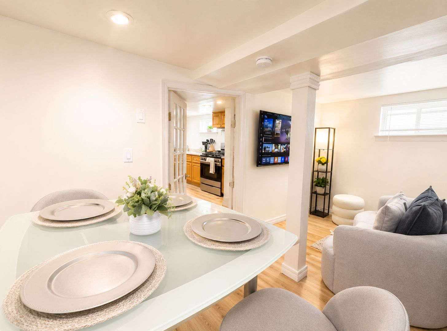 A modern dining area with a glass table set for four, a kitchen in the background, and a living area with a sofa and a TV.