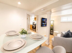 Un comedor moderno con una mesa de vidrio preparada para cuatro, una cocina al fondo y un área de estar con sofá y televisor.