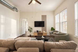 A modern living room with a sectional sofa, a coffee table, a TV mounted on the wall, and a large window with blinds. - Brand-New 2BR/2BA | Reserved Parking | Sleeps 6 (Dallas)