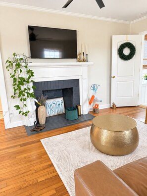 A living room with a fireplace, a television mounted above it, a door with a wreath, and a round ottoman. - Cozy 2-bedroom house in beautiful Morehead City (Morehead City)