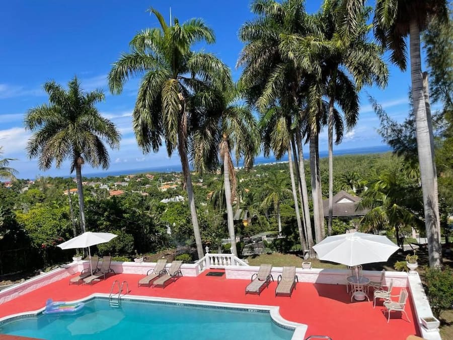 A pool area with lounge chairs and umbrellas, surrounded by palm trees and a view of the landscape.