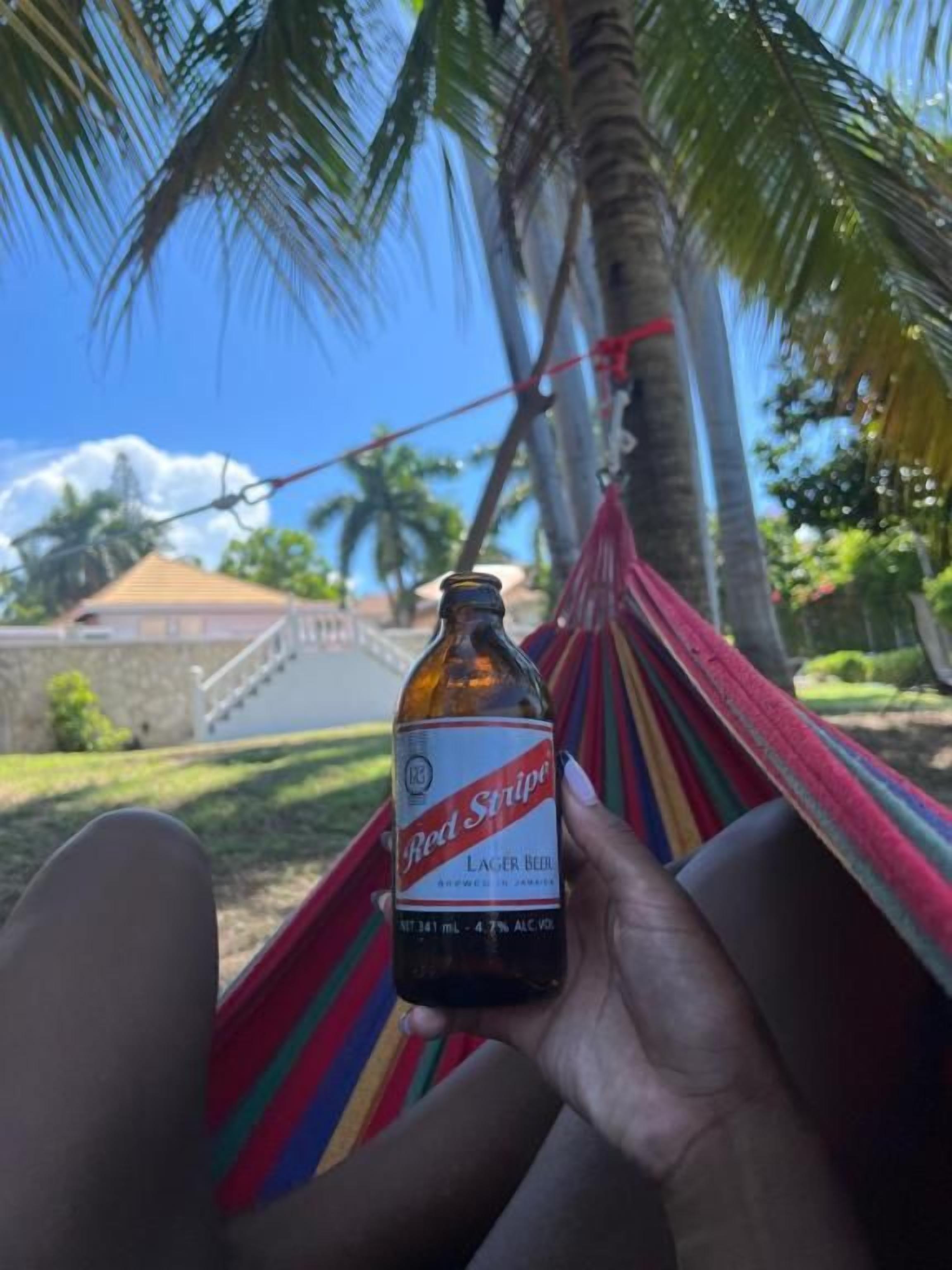 Una persona sosteniendo una botella de cerveza Red Stripe, con una hamaca y palmeras al fondo.