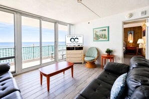 A spacious living area with a view of the ocean, a wooden table, a cushioned sofa, a wicker chair, and a small side table with a lamp. - Carousel 606 (Ocean City)