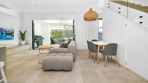 Ein modernes Wohnzimmer mit einer Couch, einem Esstisch mit Stühlen, einer Hängelampe und Blick auf einen Balkon mit Grünpflanzen.