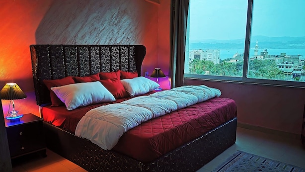 Un dormitorio con una cama grande, una mesita de noche con lámpara, vistas de la ciudad a través de una ventana y una alfombra estampada en el suelo.