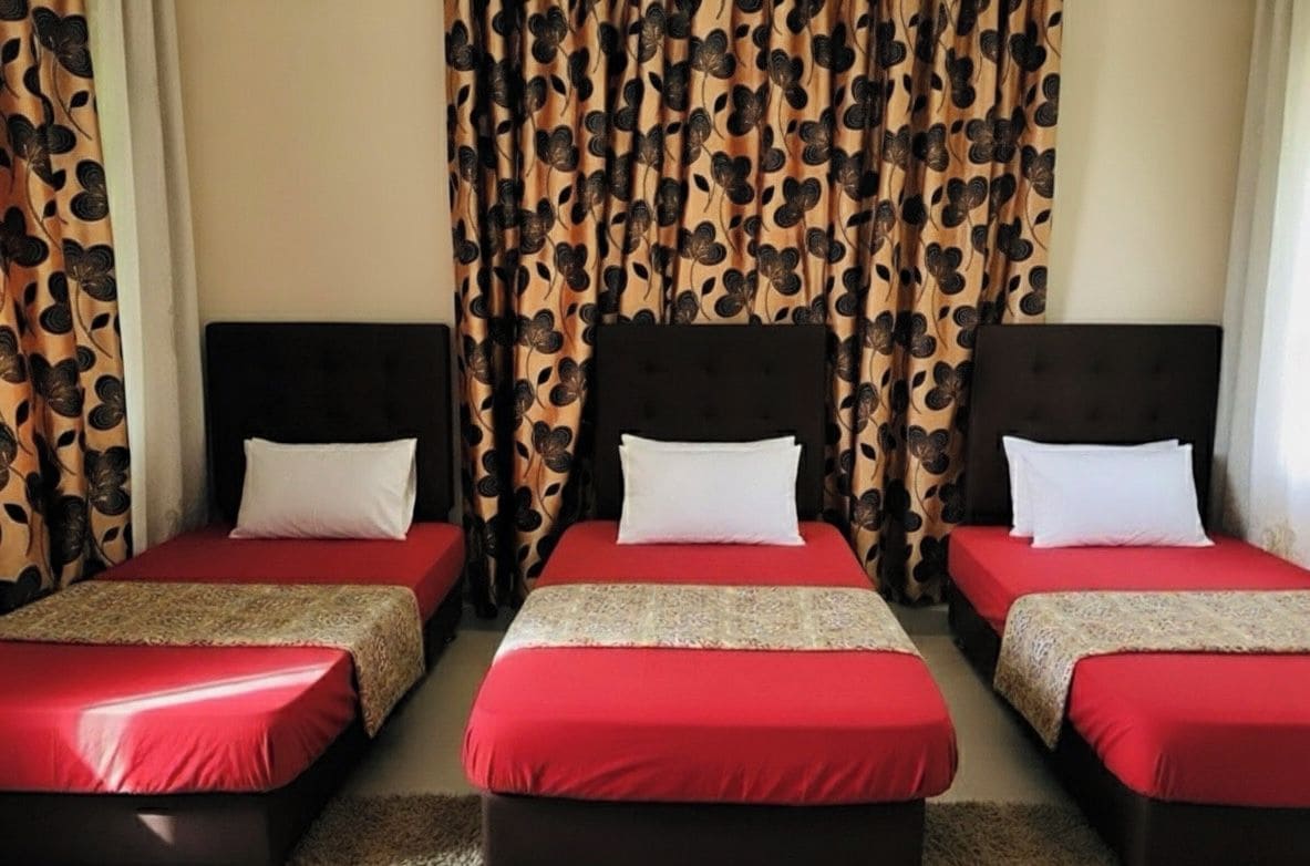 A hotel room with three single beds, each with a red bedspread and white pillows. The room has a dark headboard and a curtain with a leaf pattern.
