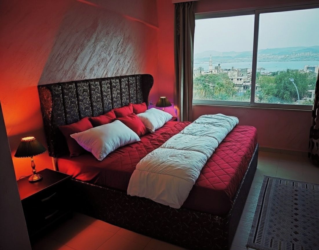 Un dormitorio con una cama grande, una mesita de noche, una lámpara y vistas a un paisaje urbano a través de una ventana.