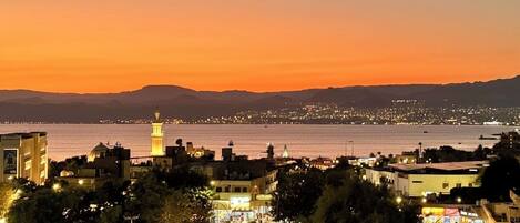 Un paisaje urbano costero al atardecer con una torre destacada, calles iluminadas y vista al mar.