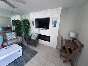 A living room with a flat-screen TV mounted on a white wall, a brown leather armchair, a wooden dining table with a lamp, and a grey sofa with floral cushions. - 2 Bedroom Siesta Key Condo Private Beach Access, Pool & Tiki Bar (Sarasota)