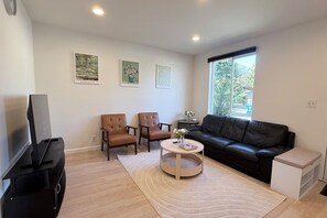 Sala de estar com sofá de couro preto, duas poltronas marrons, mesa de centro redonda, TV de tela plana e janela com vista para área verde.