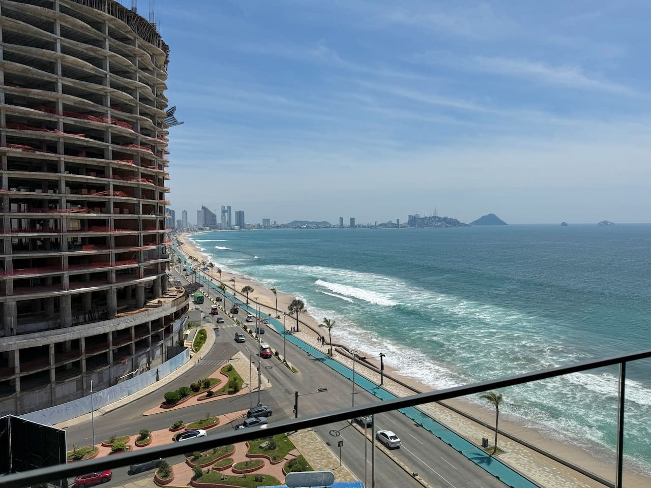 Paisagem costeira urbana com uma avenida à beira-mar, um grande edifício em construção e um horizonte distante.